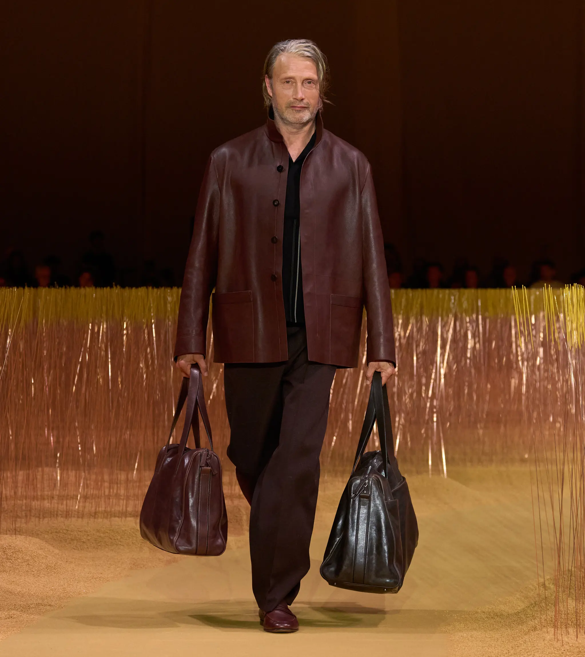 在 ZEGNA 2025 年夏季時裝表演天橋上，Mads Mikkelsen 身穿酒紅色皮䄛和深色長褲，搭配兩個皮革行李袋。